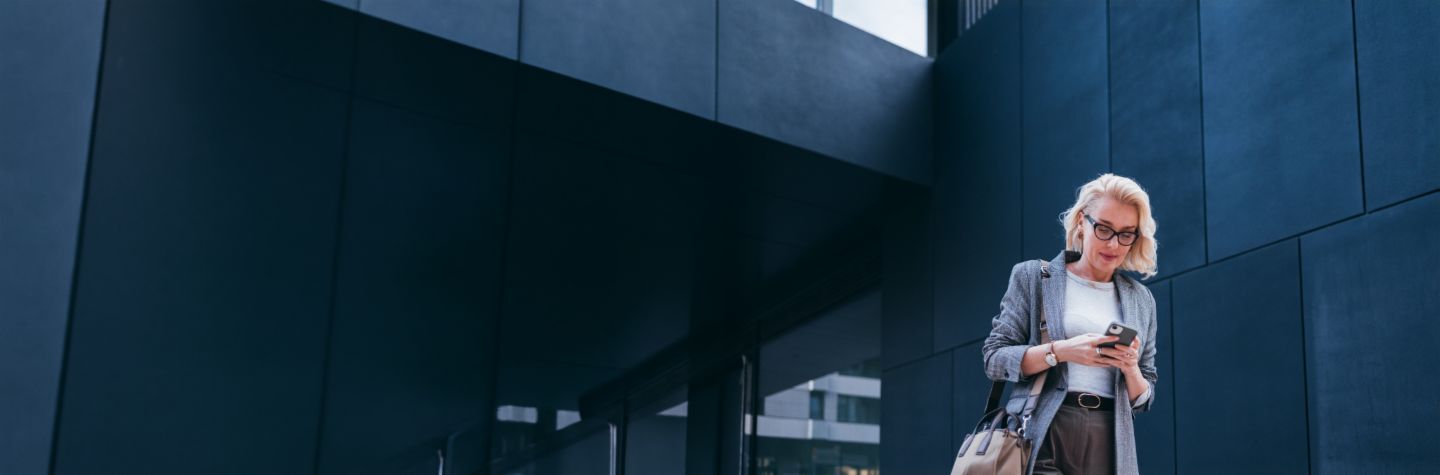Uma empresária loira de blazer cinza está em frente a um prédio moderno, olhando para seu smartphone e sorrindo.