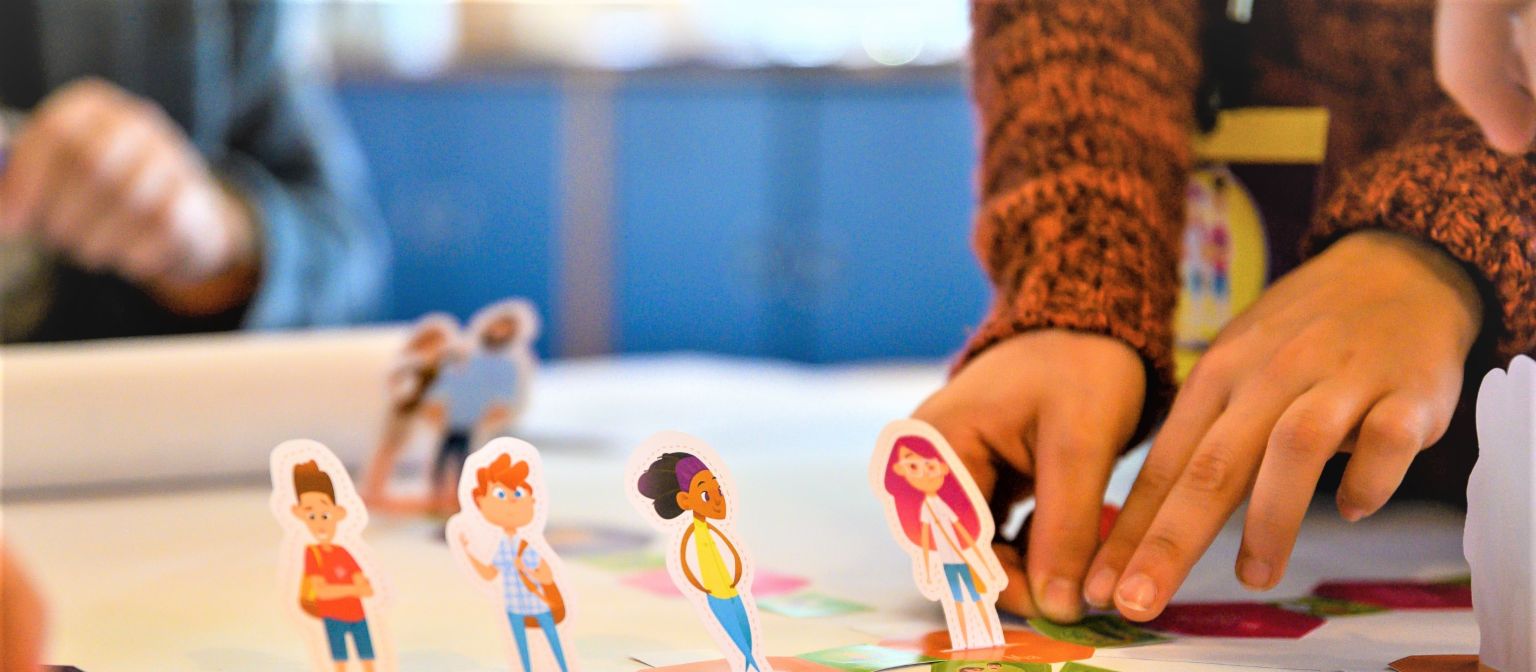 Uma foto mostra mãos movendo figuras de alunos em um tabuleiro de jogo. Abaixo, está o texto: Educação financeira: Quem aprende a lidar com o dinheiro desde cedo está melhor protegido contra o superendividamento no futuro.