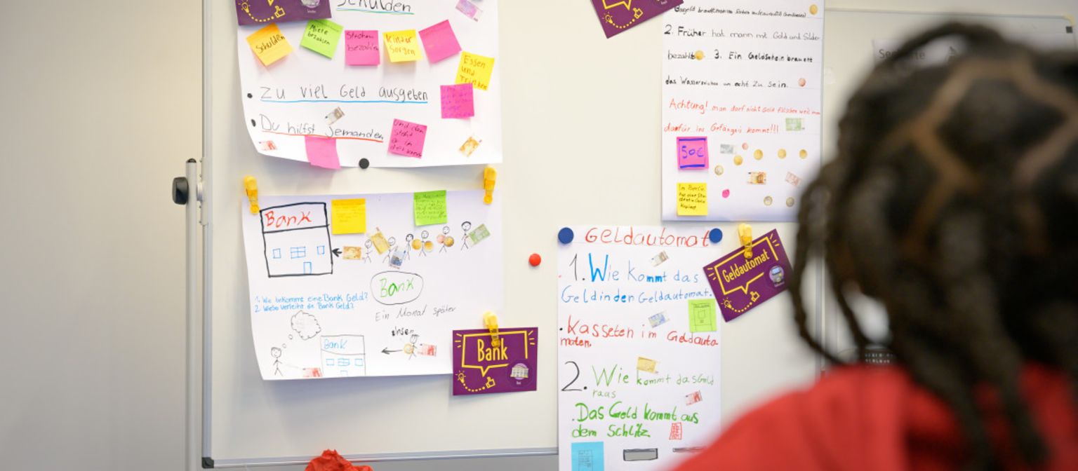 Uma foto mostra post-its e notas em um quadro, com termos como "Dívidas", "Dinheiro", "Banco" e "Caixa Eletrônico". Abaixo, está o texto: "No programa ManoMoneta da finlit foundation, pertencente à EOS, alunos com idades entre 9 e 13 anos podem adquirir conhecimentos sobre produtos financeiros."