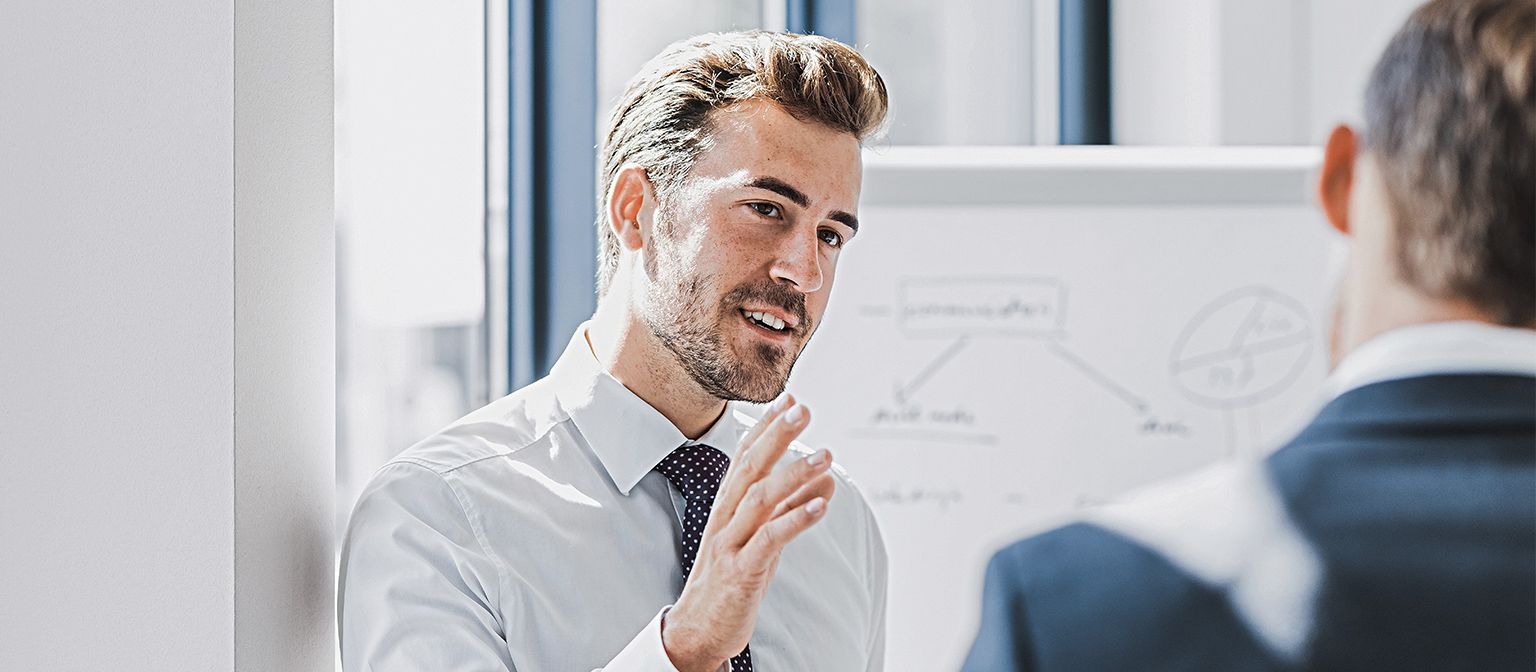 Vê-se um homem em traje profissional a explicar algo com uma expressão simpática e empenhada. À sua frente está outra pessoa que claramente o escuta. Ao fundo, vê-se um quadro branco com um esboço.
