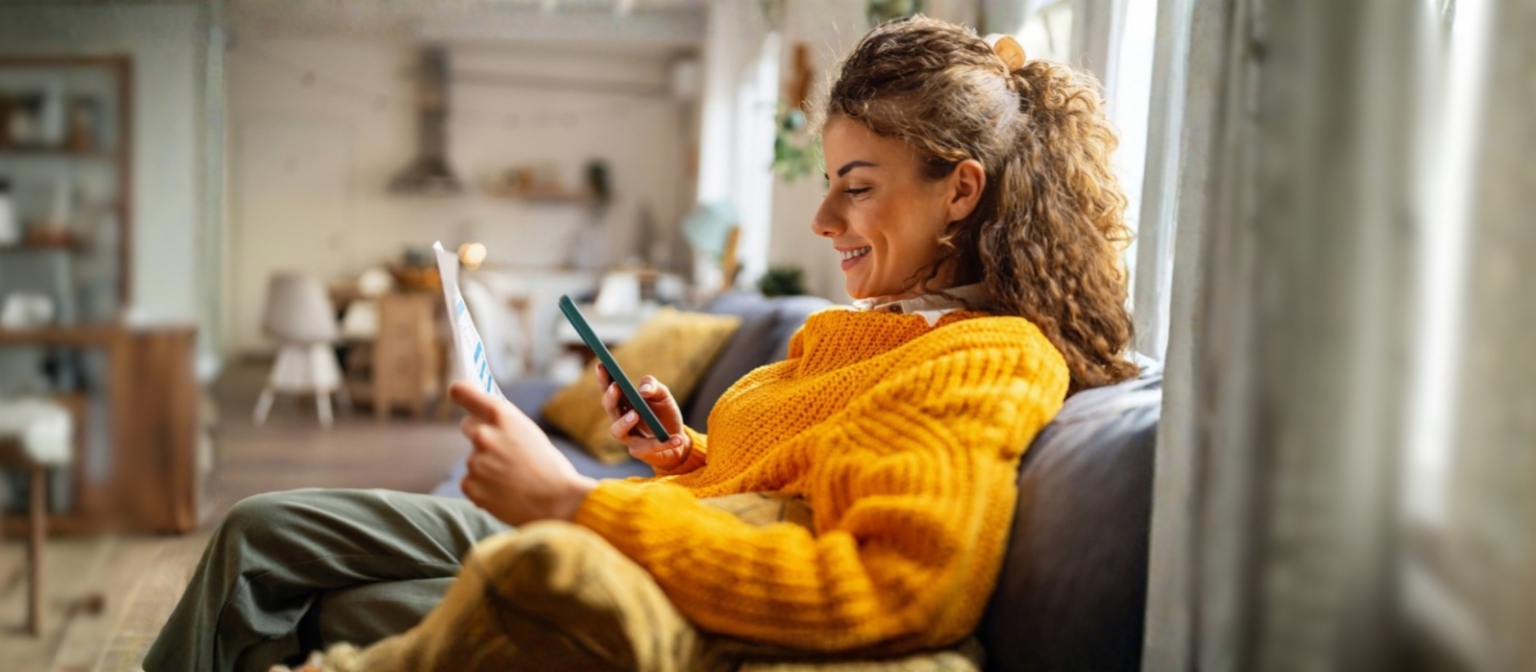 Uma jovem sorridente com cabelo cacheado, vestindo um suéter amarelo aconchegante, senta-se em um sofá na sua sala de estar e olha para o seu smartphone enquanto segura um documento de papel.