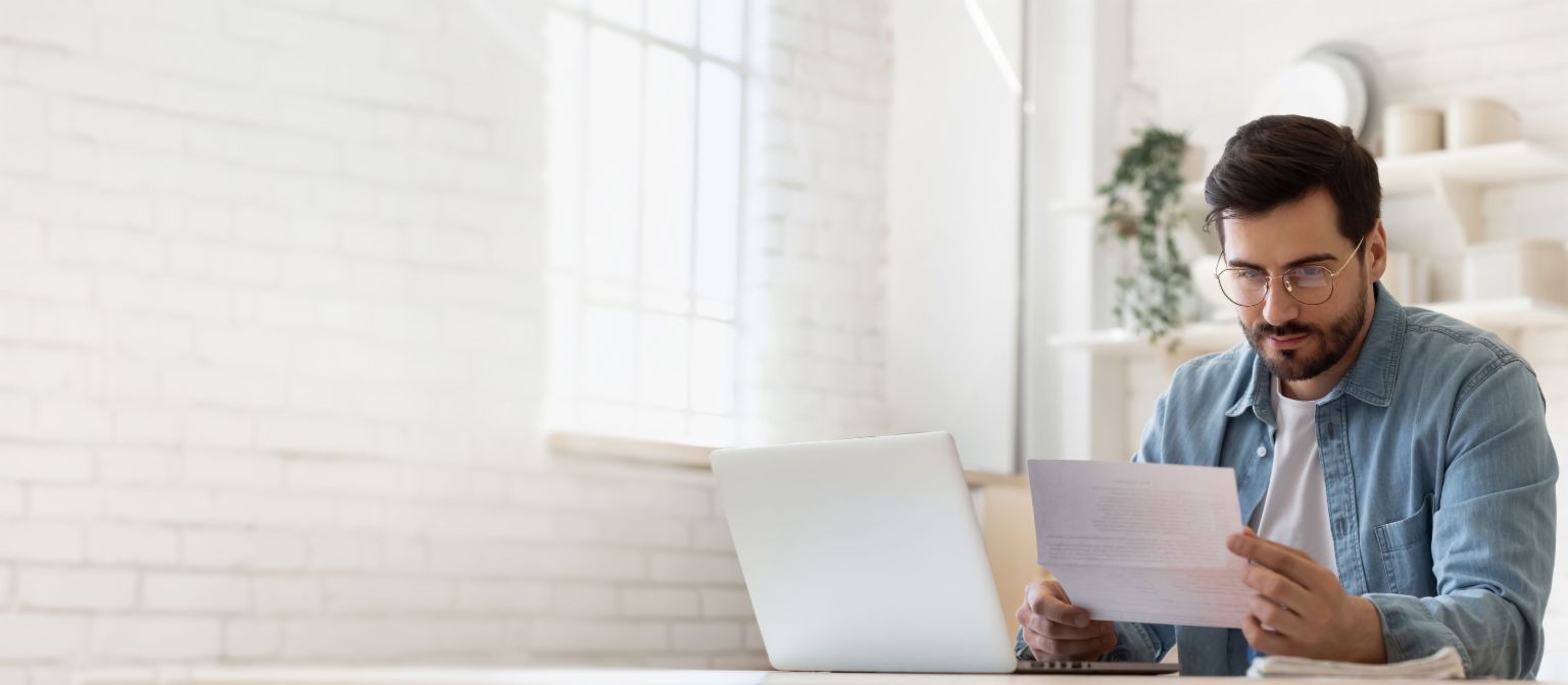 Homem de óculos trabalhando no laptop enquanto revisa documento de liquidação de dívidas em casa iluminada