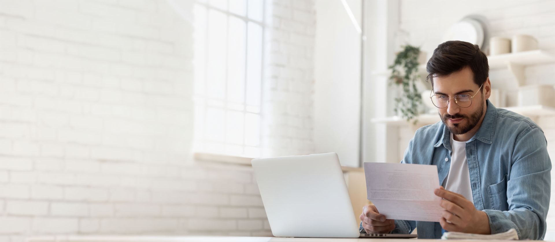 Homem de óculos trabalhando no laptop enquanto revisa documento de liquidação de dívidas em casa iluminada
