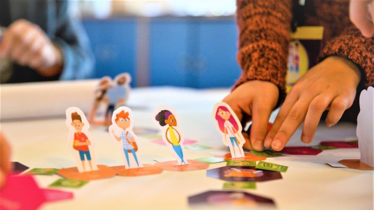 Uma foto mostra mãos movendo figuras de alunos em um tabuleiro de jogo. Abaixo, está o texto: Educação financeira: Quem aprende a lidar com o dinheiro desde cedo está melhor protegido contra o superendividamento no futuro.