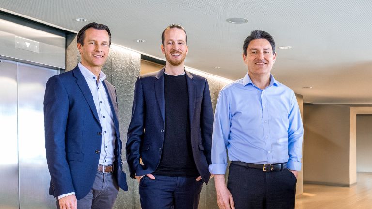 Três membros masculinos do Conselho de Administração do Grupo EOS estão de pé, vestidos com trajes de negócios, em frente a elevadores em um ambiente moderno.