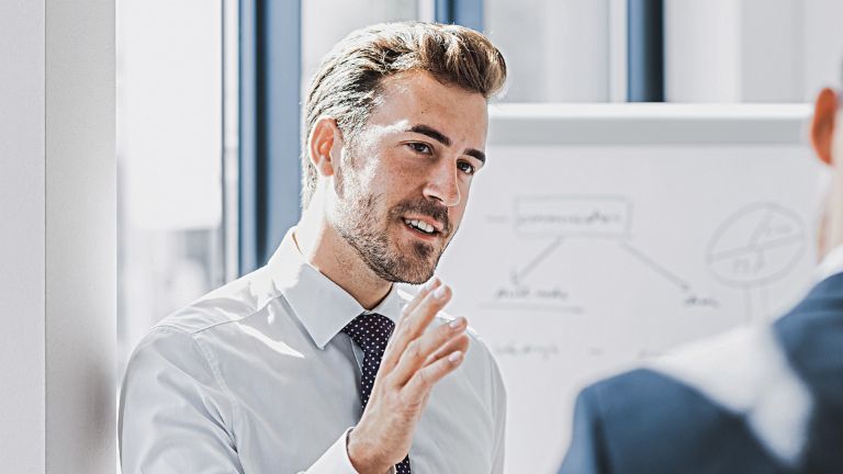 Vê-se um homem em traje profissional a explicar algo com uma expressão simpática e empenhada. À sua frente está outra pessoa que claramente o escuta. Ao fundo, vê-se um quadro branco com um esboço.