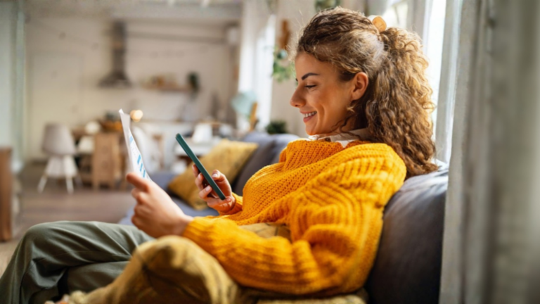 Uma jovem sorridente com cabelo cacheado, vestindo um suéter amarelo aconchegante, senta-se em um sofá na sua sala de estar e olha para o seu smartphone enquanto segura um documento de papel.
