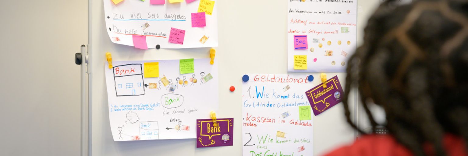 Uma foto mostra post-its e notas em um quadro, com termos como "Dívidas", "Dinheiro", "Banco" e "Caixa Eletrônico". Abaixo, está o texto: "No programa ManoMoneta da finlit foundation, pertencente à EOS, alunos com idades entre 9 e 13 anos podem adquirir conhecimentos sobre produtos financeiros."