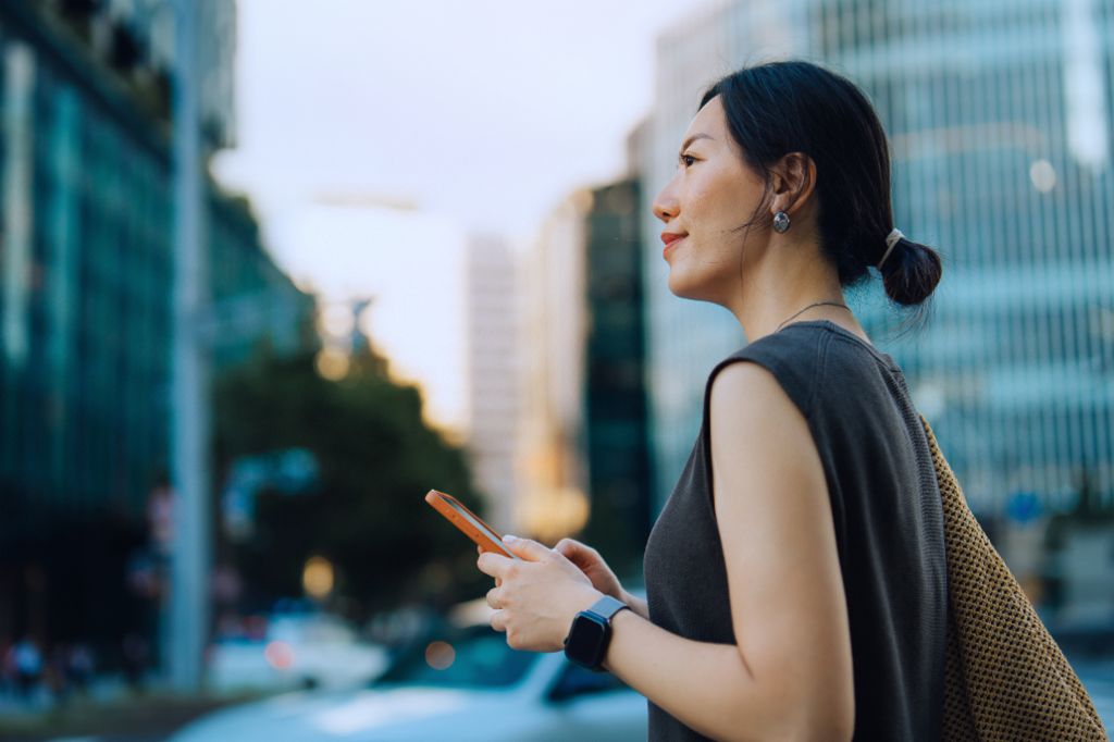 Uma jovem mulher está ao ar livre em um ambiente urbano, segurando seu smartphone e sorrindo enquanto olha para frente. Ao fundo, há prédios modernos e árvores.