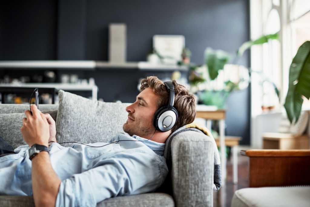 Um jovem está deitado num sofá na sua sala de estar, a usar auscultadores e a olhar para o seu smartphone enquanto sorri, a desfrutar de música ou de um podcast.