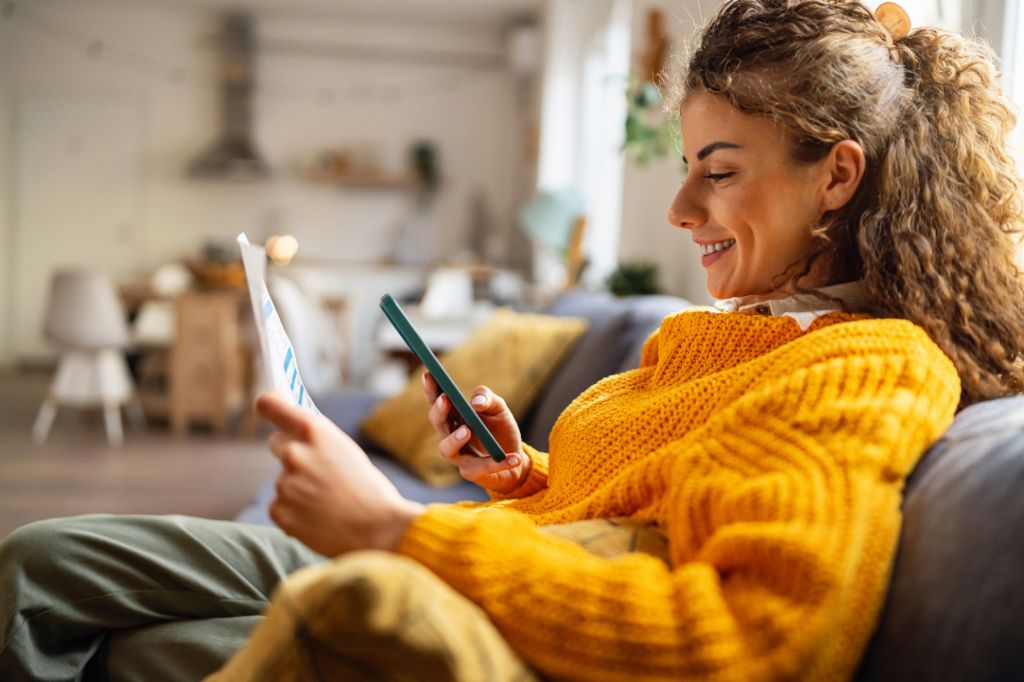 Uma jovem sorridente com cabelo cacheado, vestindo um suéter amarelo aconchegante, senta-se em um sofá na sua sala de estar e olha para o seu smartphone enquanto segura um documento de papel.