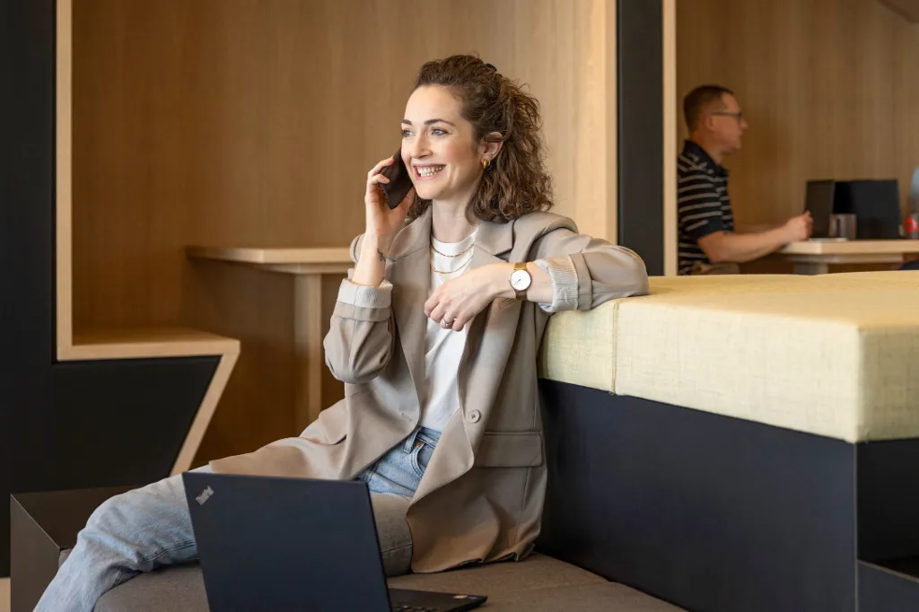 Uma pessoa sorridente está sentada numa moderna zona lounge de escritório, a falar ao telefone e a trabalhar num portátil. Ao fundo, duas outras pessoas estão a trabalhar.