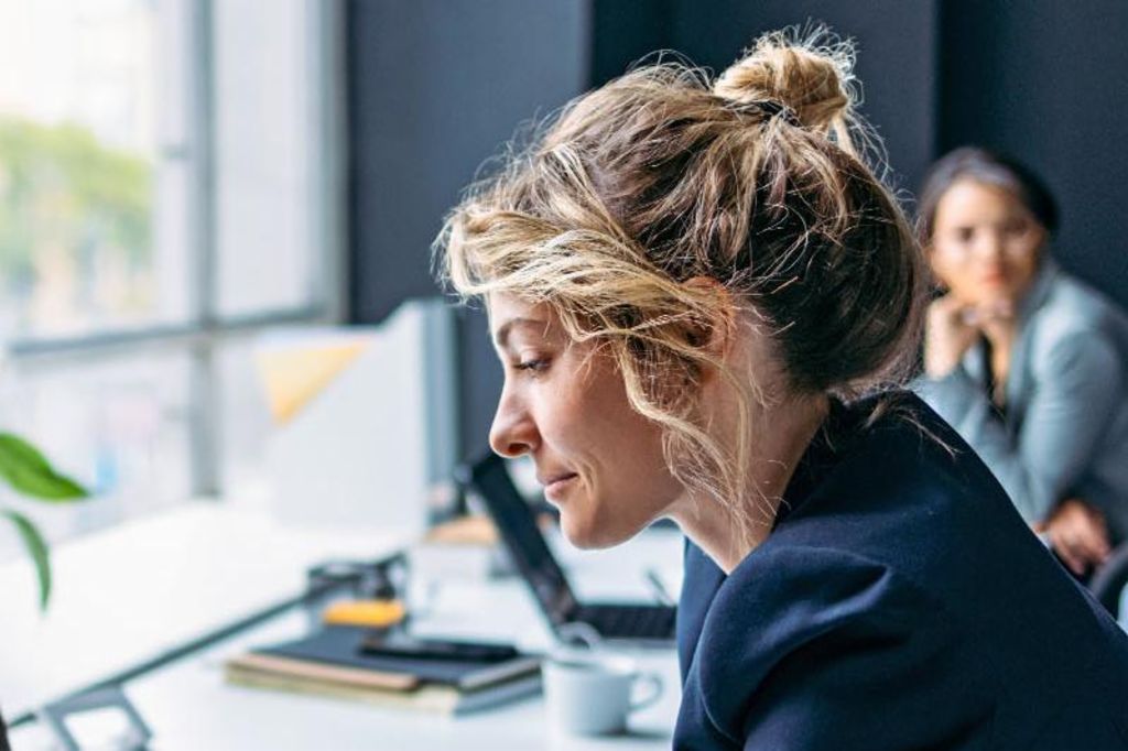A imagem mostra uma mulher sentada em uma mesa em um ambiente de escritório moderno, vestindo um blazer escuro enquanto se concentra em seu laptop, com uma planta e alguns suprimentos de escritório visíveis na mesa, e outra pessoa sentada ao fundo.