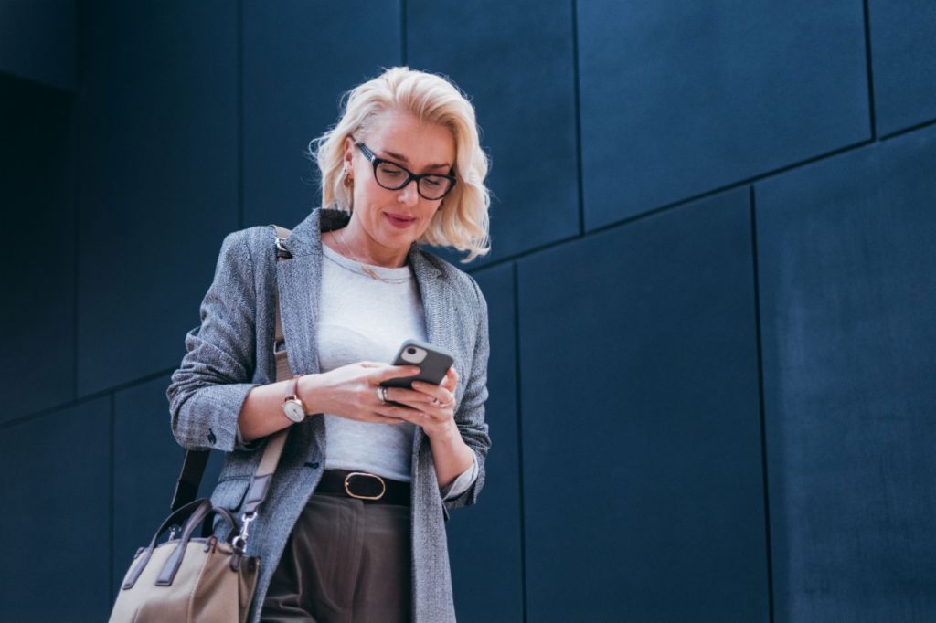 Uma empresária loira de blazer cinza está em frente a um prédio moderno, olhando para seu smartphone e sorrindo.
