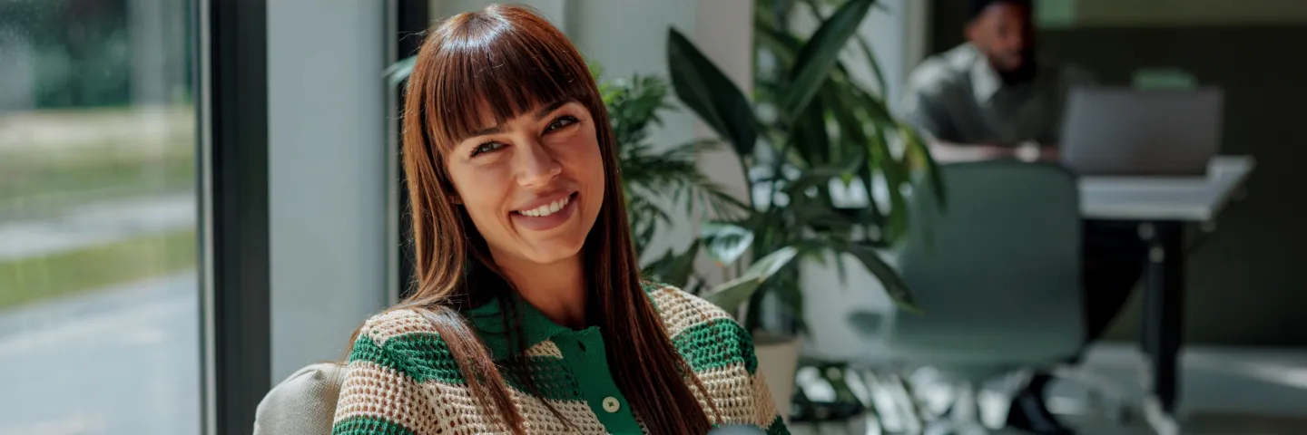 Uma mulher sorridente de cabelo escuro está sentada confortavelmente num escritório luminoso e moderno e olha de forma calorosa para a câmara. Ao fundo, veem-se outras pessoas e grandes janelas.