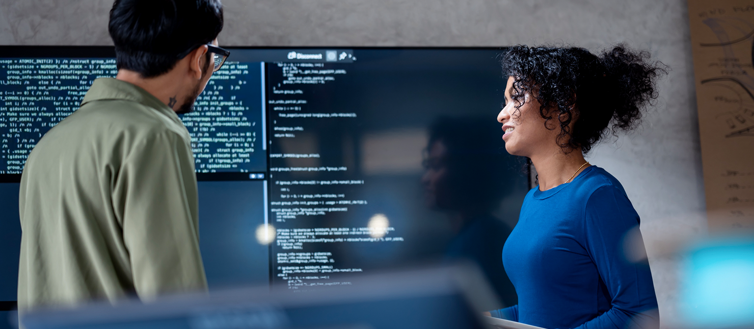 IA na recuperação de dívidas: Nesta imagem, dois profissionais estão analisando colaborativamente o código do programa para processos automatizados em uma grande tela. A cena ilustra a aplicação prática da Inteligência Artificial (IA) e da Automação de Processos Robóticos (RPA) na gestão de dívidas — um exemplo de transformação digital na gestão de recebíveis.