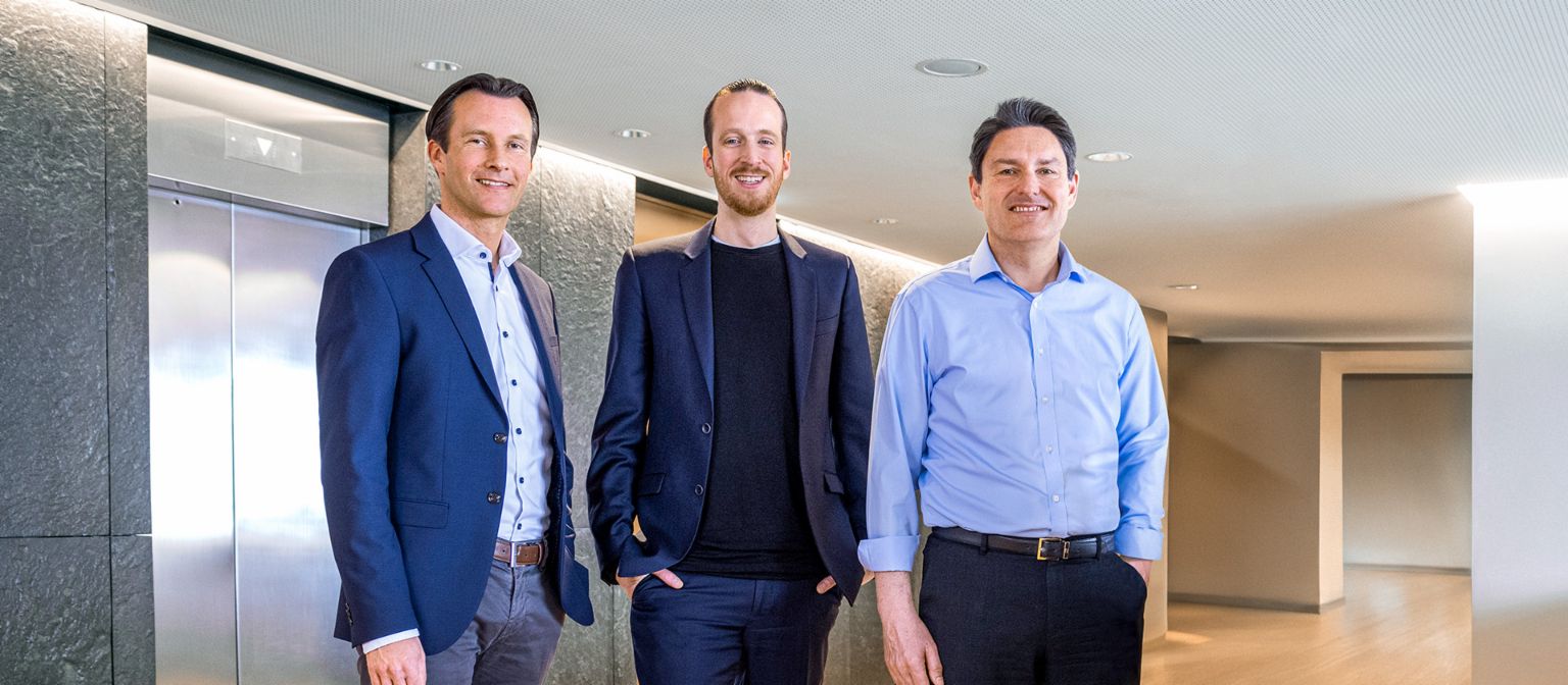 Três membros masculinos do Conselho de Administração do Grupo EOS estão de pé, vestidos com trajes de negócios, em frente a elevadores em um ambiente moderno.
