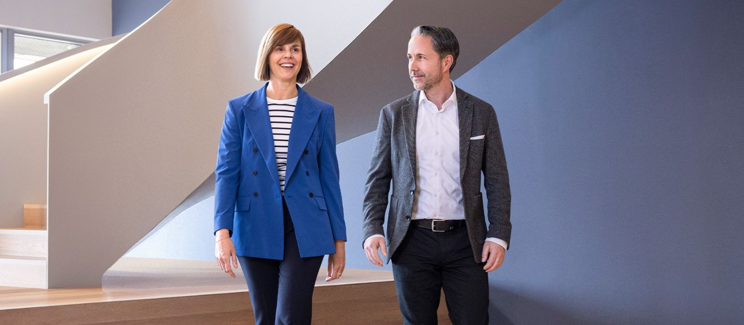 O CEO em um blazer cinza e a CFO do Grupo EOS em um blazer azul caminham por um escritório moderno. O espaço conta com grandes janelas, uma parede curva e elegantes lâmpadas pendentes.
