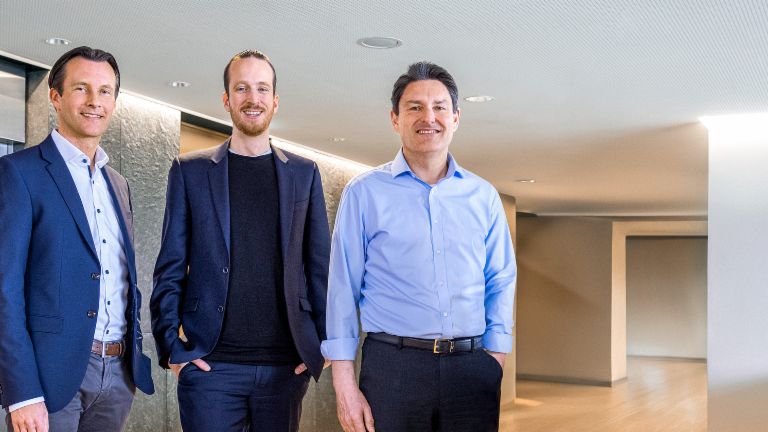 Três membros masculinos do Conselho de Administração do Grupo EOS estão de pé, vestidos com trajes de negócios, em frente a elevadores em um ambiente moderno.
