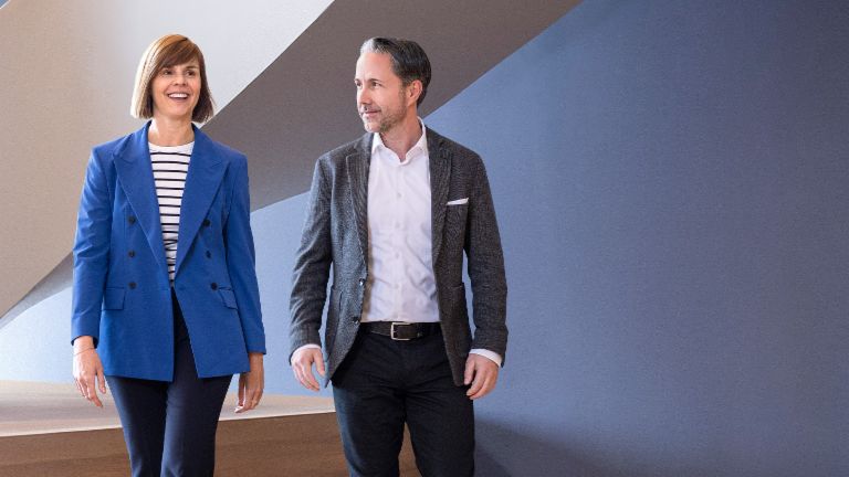 O CEO em um blazer cinza e a CFO do Grupo EOS em um blazer azul caminham por um escritório moderno. O espaço conta com grandes janelas, uma parede curva e elegantes lâmpadas pendentes.