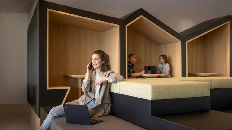 Uma pessoa sorridente está sentada numa moderna zona lounge de escritório, a falar ao telefone e a trabalhar num portátil. Ao fundo, duas outras pessoas estão a trabalhar.
