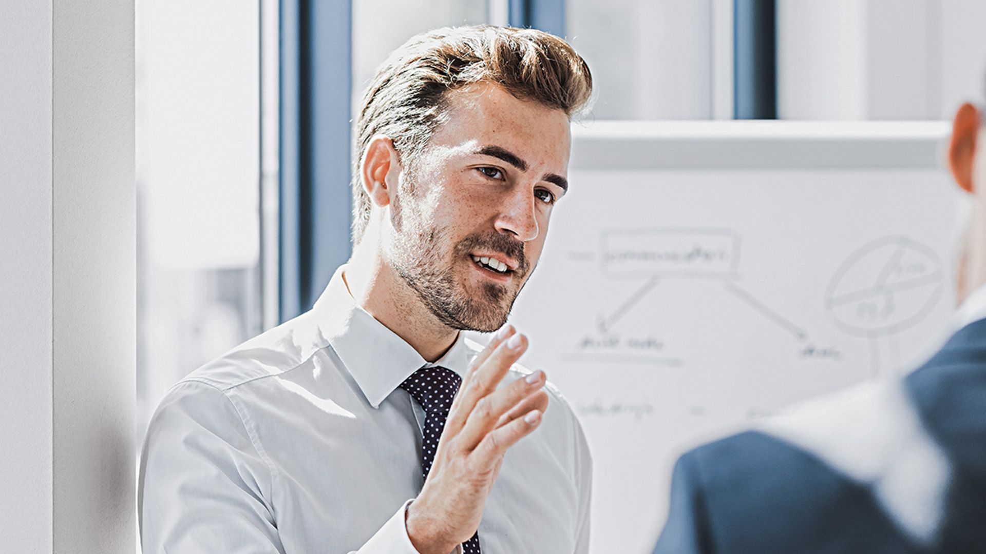 Vê-se um homem em traje profissional a explicar algo com uma expressão simpática e empenhada. À sua frente está outra pessoa que claramente o escuta. Ao fundo, vê-se um quadro branco com um esboço.