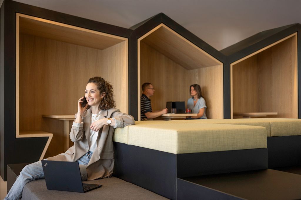 Uma pessoa sorridente está sentada numa moderna zona lounge de escritório, a falar ao telefone e a trabalhar num portátil. Ao fundo, duas outras pessoas estão a trabalhar.