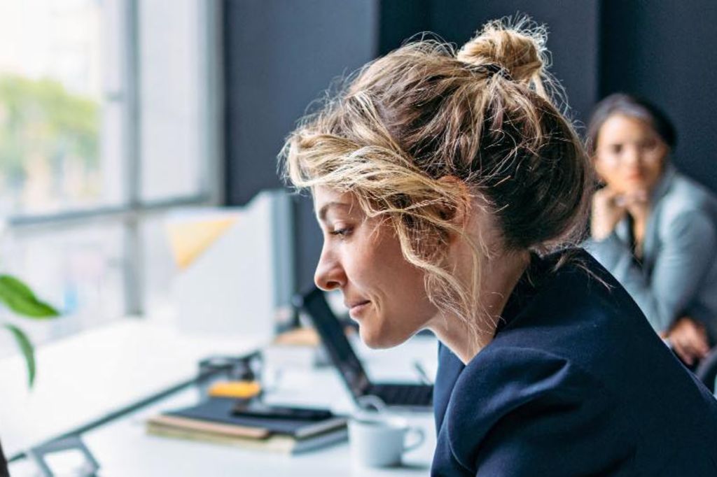 A imagem mostra uma mulher sentada em uma mesa em um ambiente de escritório moderno, vestindo um blazer escuro enquanto se concentra em seu laptop, com uma planta e alguns suprimentos de escritório visíveis na mesa, e outra pessoa sentada ao fundo.