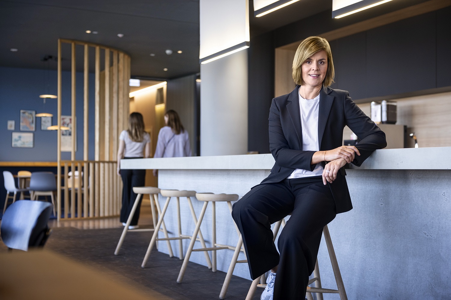 Dr. Eva Griewel, CFO do Grupo EOS, está sentada em um espaço social em um banquinho alto, vestindo um blazer preto.