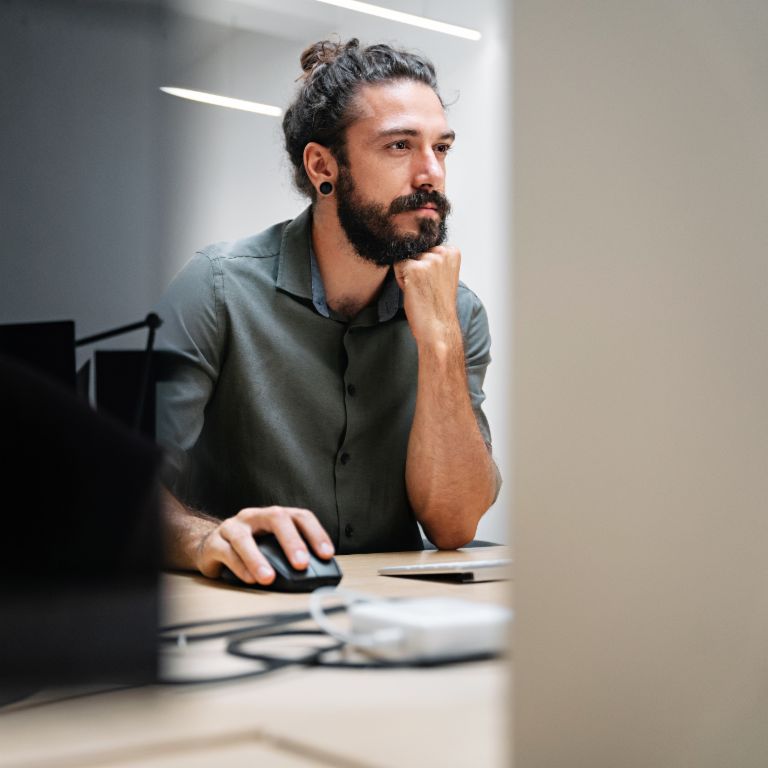 Um homem barbudo com cabelo longo preso em um coque está sentado em um computador, totalmente concentrado em seu trabalho.