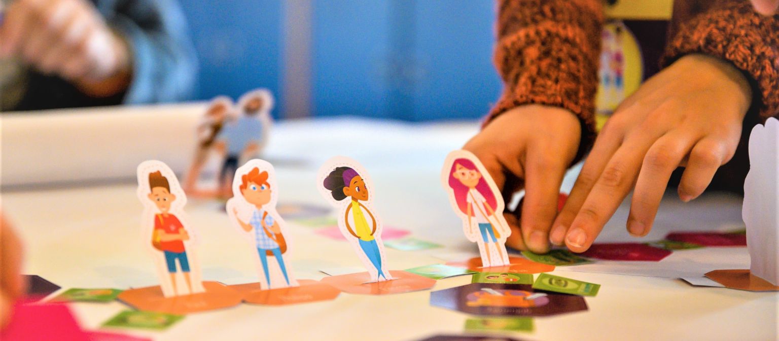 Uma foto mostra mãos movendo figuras de alunos em um tabuleiro de jogo. Abaixo, está o texto: Educação financeira: Quem aprende a lidar com o dinheiro desde cedo está melhor protegido contra o superendividamento no futuro.
