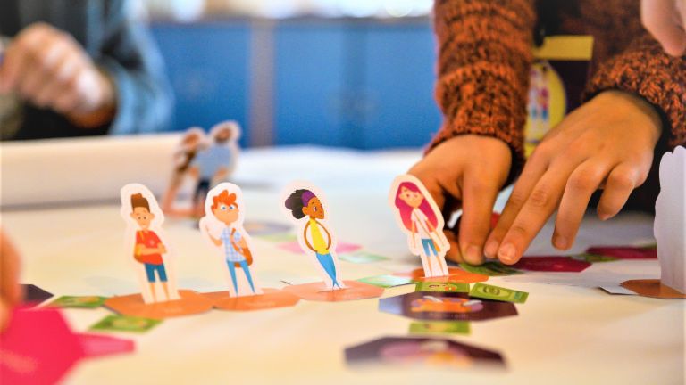 Uma foto mostra mãos movendo figuras de alunos em um tabuleiro de jogo. Abaixo, está o texto: Educação financeira: Quem aprende a lidar com o dinheiro desde cedo está melhor protegido contra o superendividamento no futuro.