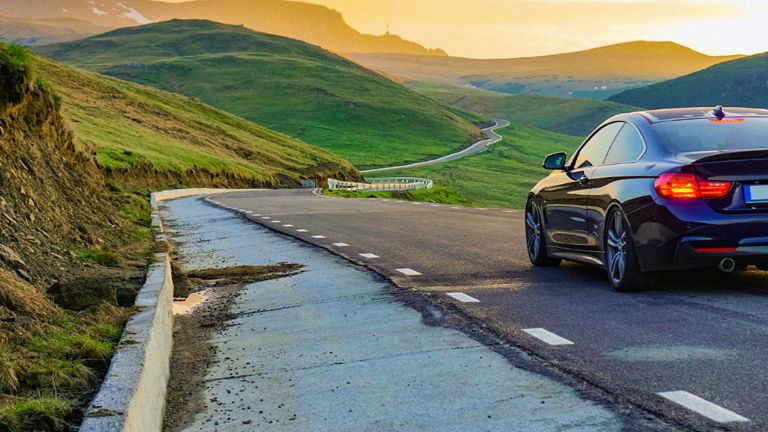 A photomontage shows a car traveling toward the sunset in the right lane of a paved road in an open, green landscape. The asphalt on the opposite lane is completely worn down.
