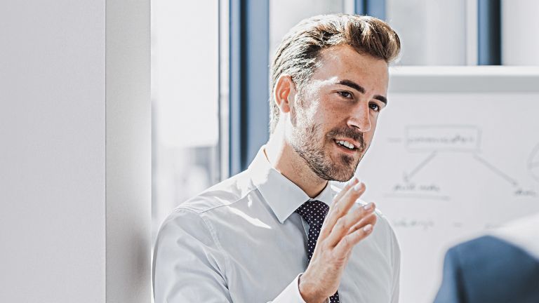 Vê-se um homem em traje profissional a explicar algo com uma expressão simpática e empenhada. À sua frente está outra pessoa que claramente o escuta. Ao fundo, vê-se um quadro branco com um esboço.