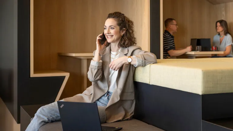 Uma pessoa sorridente está sentada numa moderna zona lounge de escritório, a falar ao telefone e a trabalhar num portátil. Ao fundo, duas outras pessoas estão a trabalhar.