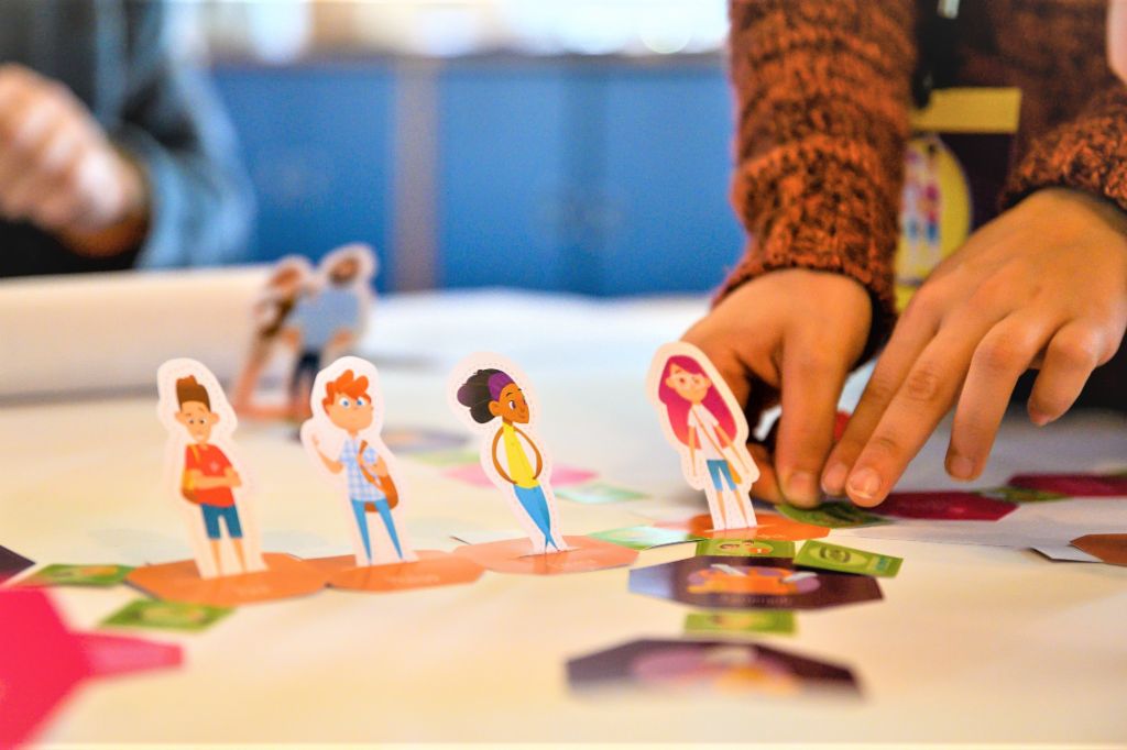 Uma foto mostra mãos movendo figuras de alunos em um tabuleiro de jogo. Abaixo, está o texto: Educação financeira: Quem aprende a lidar com o dinheiro desde cedo está melhor protegido contra o superendividamento no futuro.
