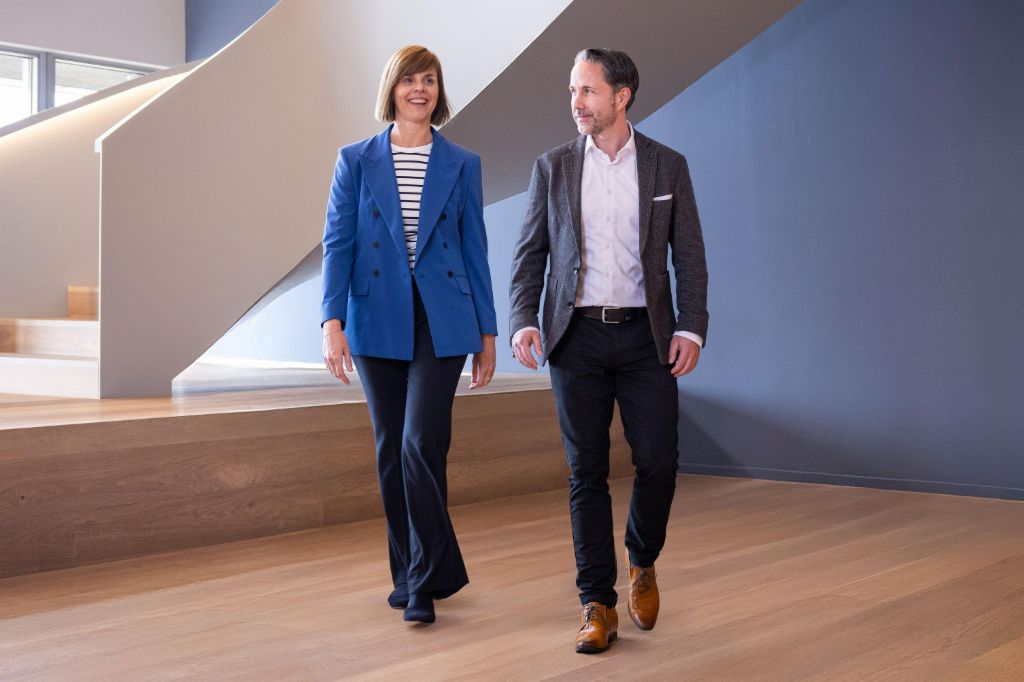 O CEO em um blazer cinza e a CFO do Grupo EOS em um blazer azul caminham por um escritório moderno. O espaço conta com grandes janelas, uma parede curva e elegantes lâmpadas pendentes.